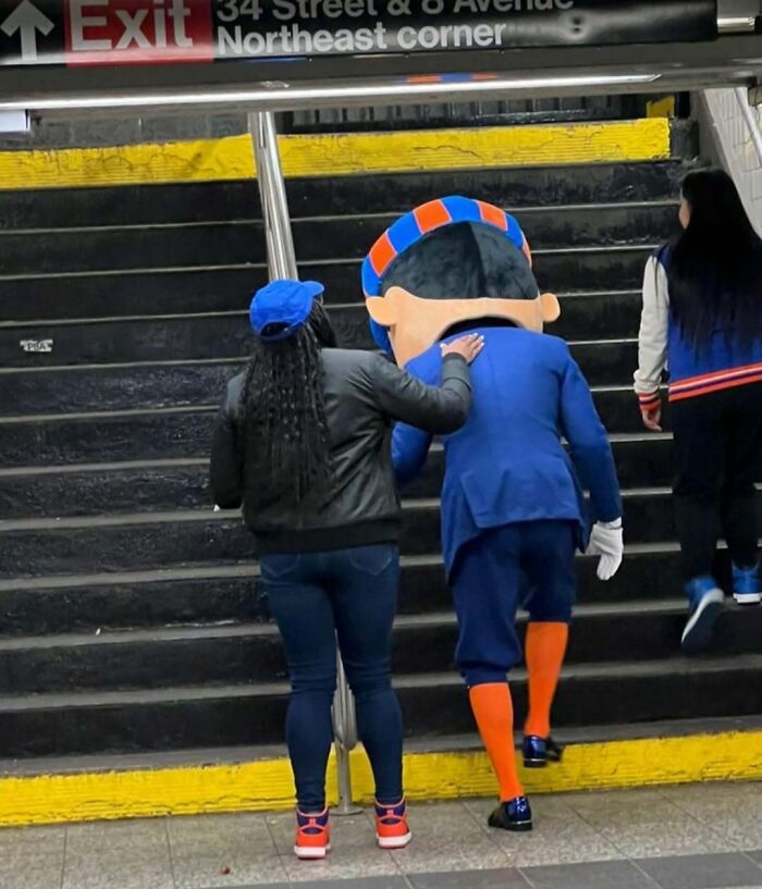 A person in a costume with a funny design being helped by a subway passenger up stairs.