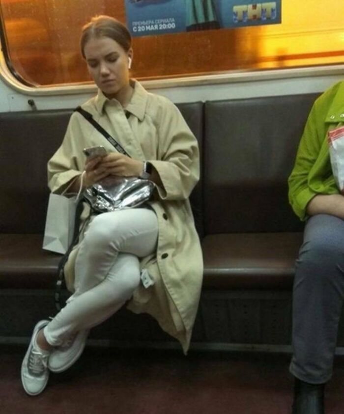 Passenger in trench coat sitting on subway train, using phone with earbuds in.