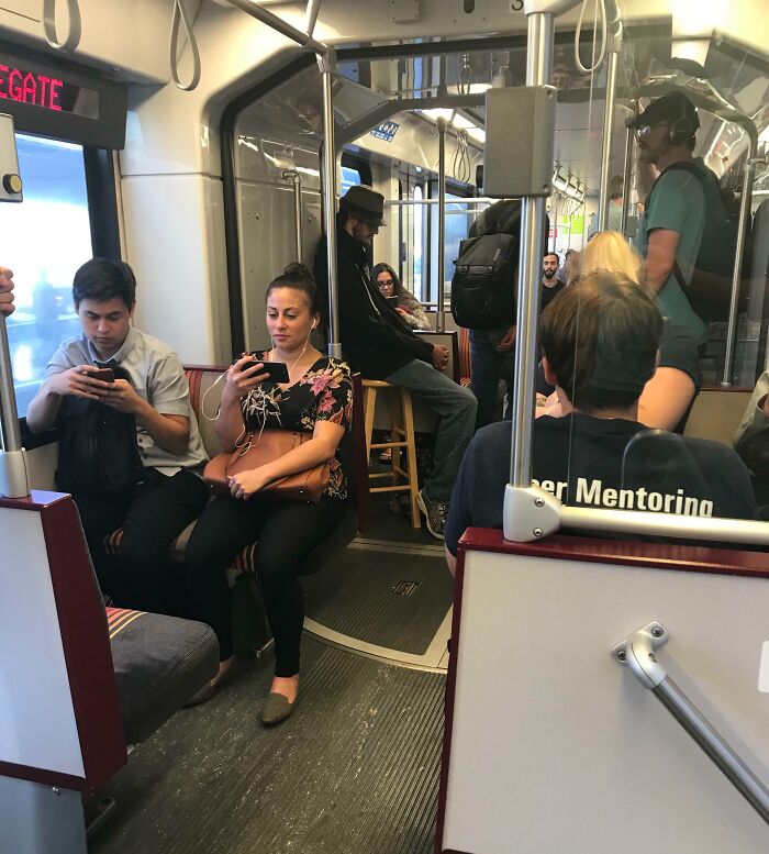 Passengers engaged with phones on a subway, one seated on a stool, creating a funny train scene.