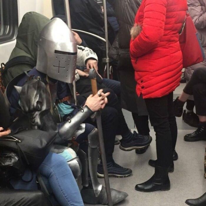 Knight in armor seated among funny subway passengers.