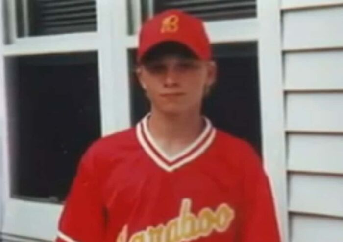 Person wearing a red baseball jersey and cap, standing in front of a window. True crime story context.