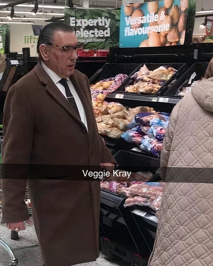 Man in a supermarket, wearing a brown coat and tie, standing beside vegetable displays. British humor caption says "Veggie Kray."