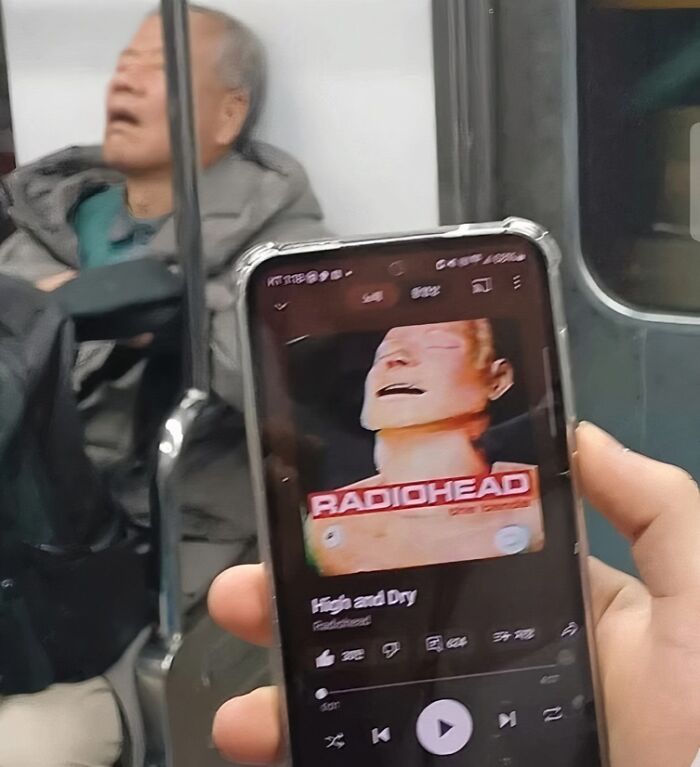 Person on subway resembles Radiohead album cover on phone screen; a humorous scene highlighting British music culture.