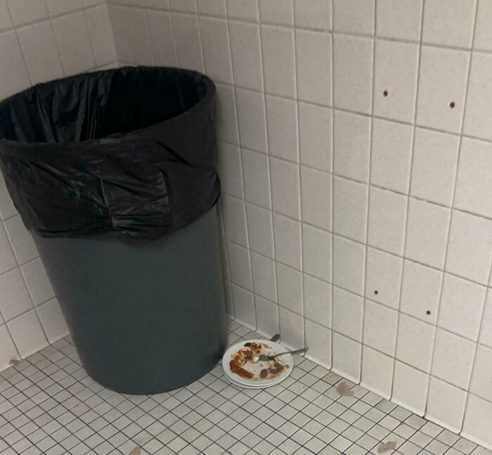 Trash can beside a tile wall with a dirty plate and fork on the floor, illustrating people being complete jerks.