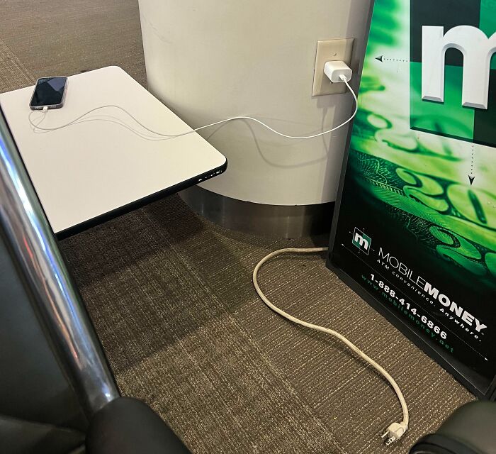 Phone charging from wall socket using a cord through an advertisement board, demonstrating people being complete jerks.