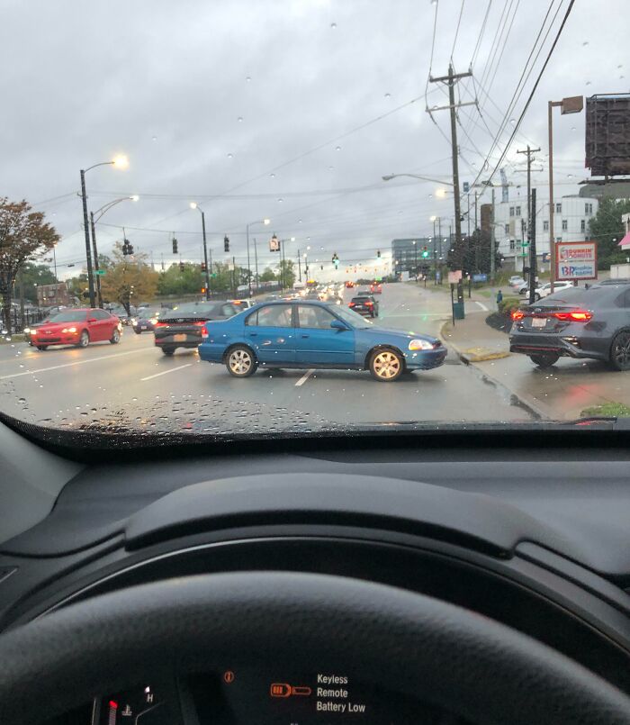 A blue car blocks an intersection, causing traffic disruption on a rainy day.