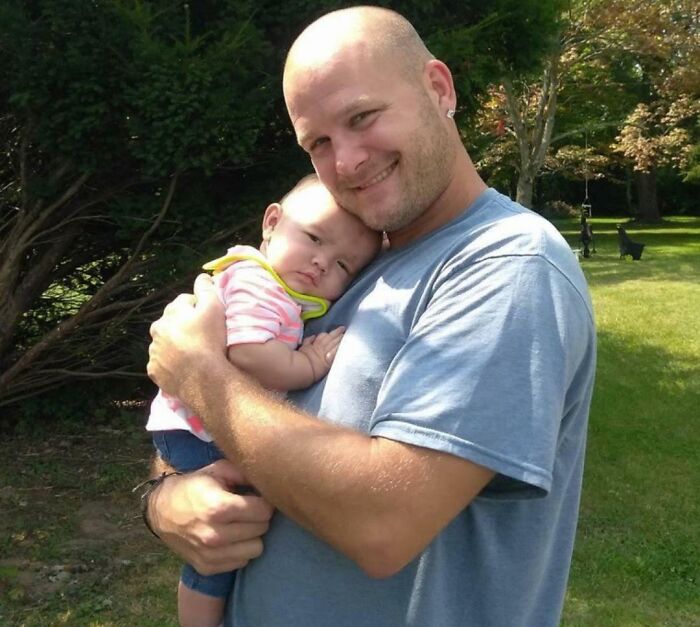 Bald man in a blue shirt holding a baby, smiling outdoors. True crime keyword image.