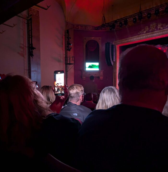 Audience in a theater with someone holding a phone high, being complete jerks by blocking the view.