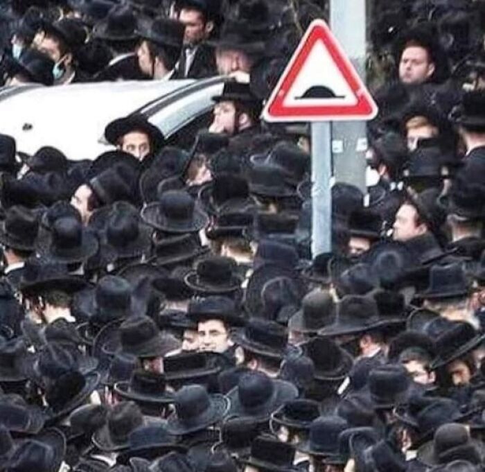 Crowded gathering of people in black hats near a road sign, featuring a bizarre scene.