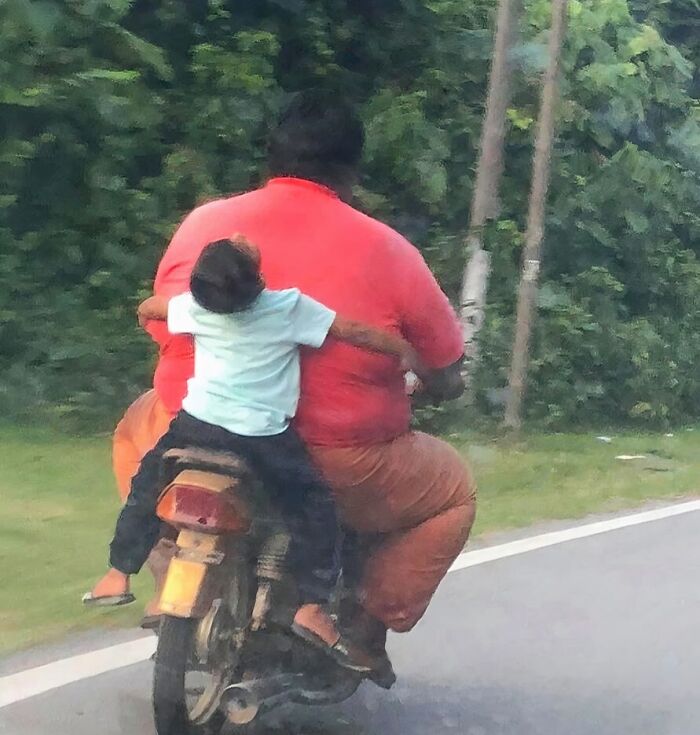 Bizarre photo of a large person on a scooter with a child clinging tightly on a road lined with greenery.