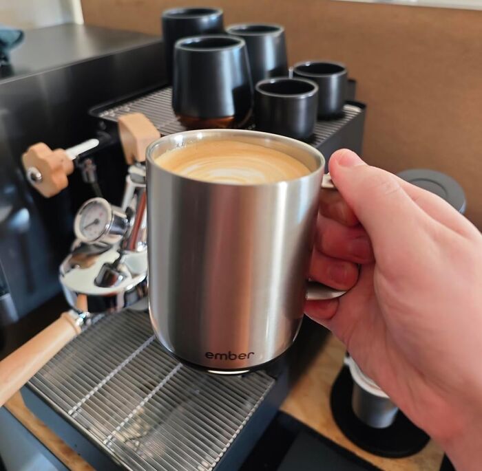 Hand holding a latte, in a silver Ember mug, next to coffee machine. Perfect gift among best Valentines Day finds.