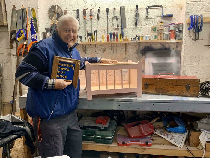 Elderly man showcasing a woodworking project of a doll-sized bed, holding a sign reading "Measure Twice, Cut Once" in a workshop.