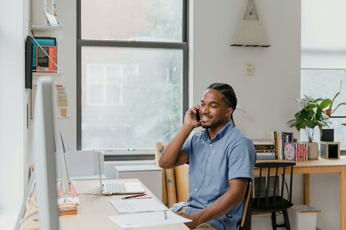 Man in blue shirt on a phone call at a desk, working on a laptop, representing unethical life hack in a modern office setting.