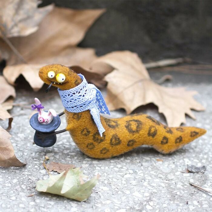 Strange art doll of a funny-looking animal with a blue scarf holding a hat, surrounded by leaves.