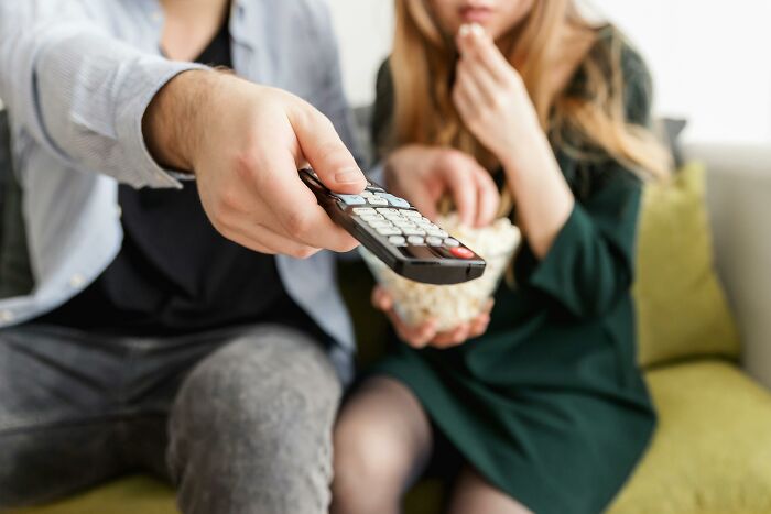 A person holding a remote control, sitting with another person who is eating popcorn, on a couch.