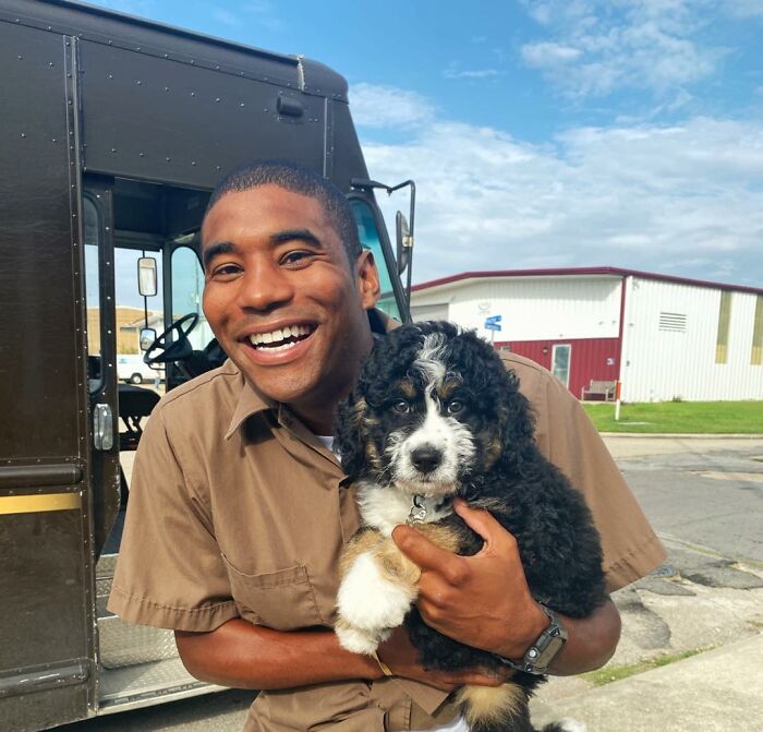 UPS driver smiling and holding an adorable puppy in front of a delivery truck.