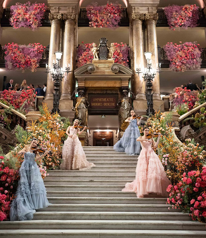 Violinists in elegant dresses perform on grand staircase at luxurious wedding venue adorned with flowers.