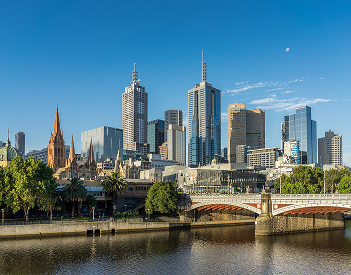 Modern city skyline with skyscrapers and historic architecture under a clear blue sky, reflecting on a calm river.