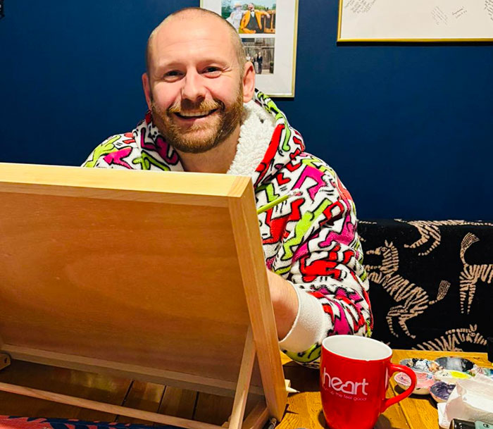 Man smiling while painting, wearing a colorful hoodie, with a red mug labeled "heart" nearby, related to viral painting.