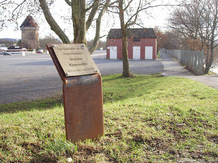 Memorial plaque in a park, linked to true crime plot twists, with buildings in the background.