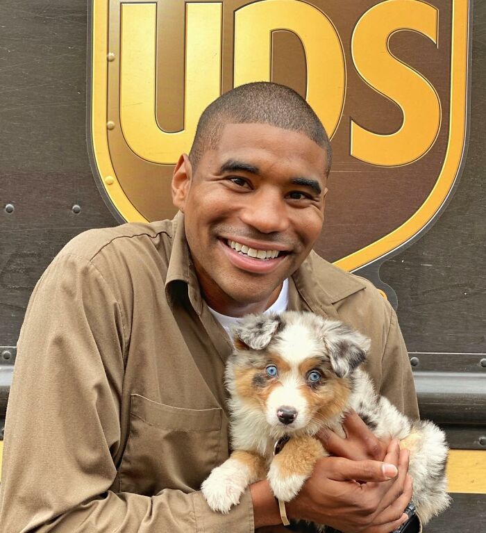 UPS driver smiling and holding an adorable puppy in front of a UPS truck.