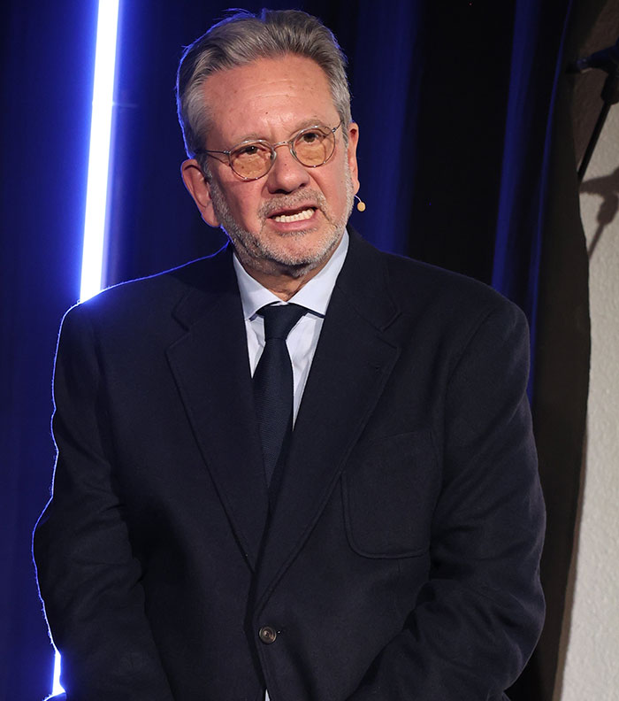 Man in a suit with glasses speaking on stage about elegance and class.