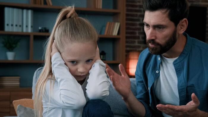 Child covering ears while sitting on a couch, as an adult gestures nearby, illustrating a messed-up situation.