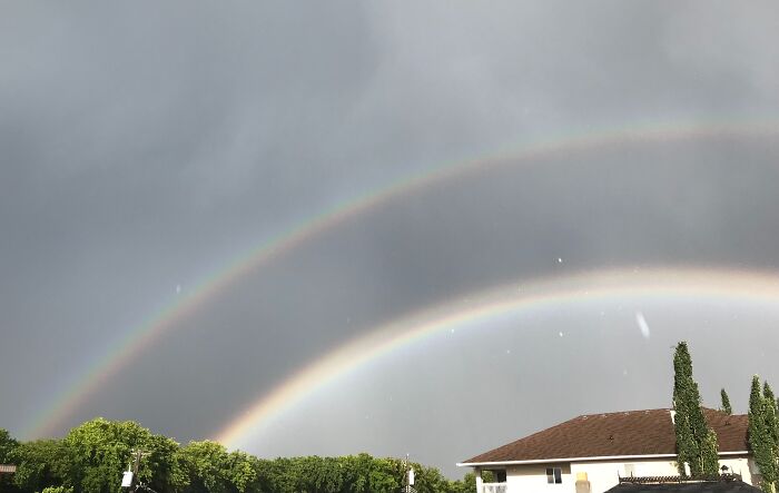 Double rainbow over a house and trees, showcasing stunning natural phenomena under a cloudy sky.