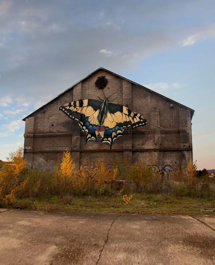 Hyper-realistic butterfly mural painted on an old brick building by a French artist, surrounded by autumn foliage.