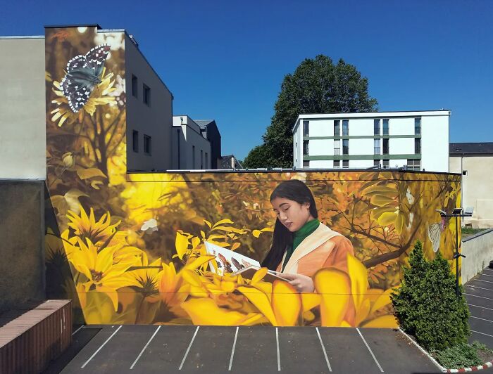 Hyper-realistic mural featuring a butterfly and a girl reading, painted on a building in a vibrant floral setting.