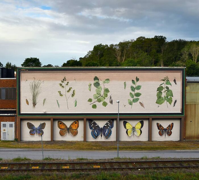 Hyper-realistic murals of butterflies and plants decorate a building wall against a backdrop of trees and cloudy sky.