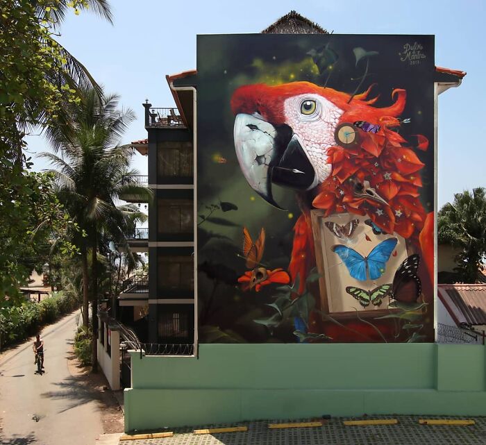 Hyper-realistic mural of a parrot with accurate butterflies painted by a French artist on a building wall.