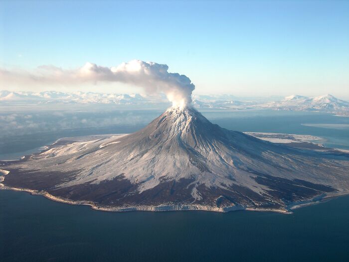 Volcano emitting smoke surrounded by ocean, showcasing stunning natural phenomena.