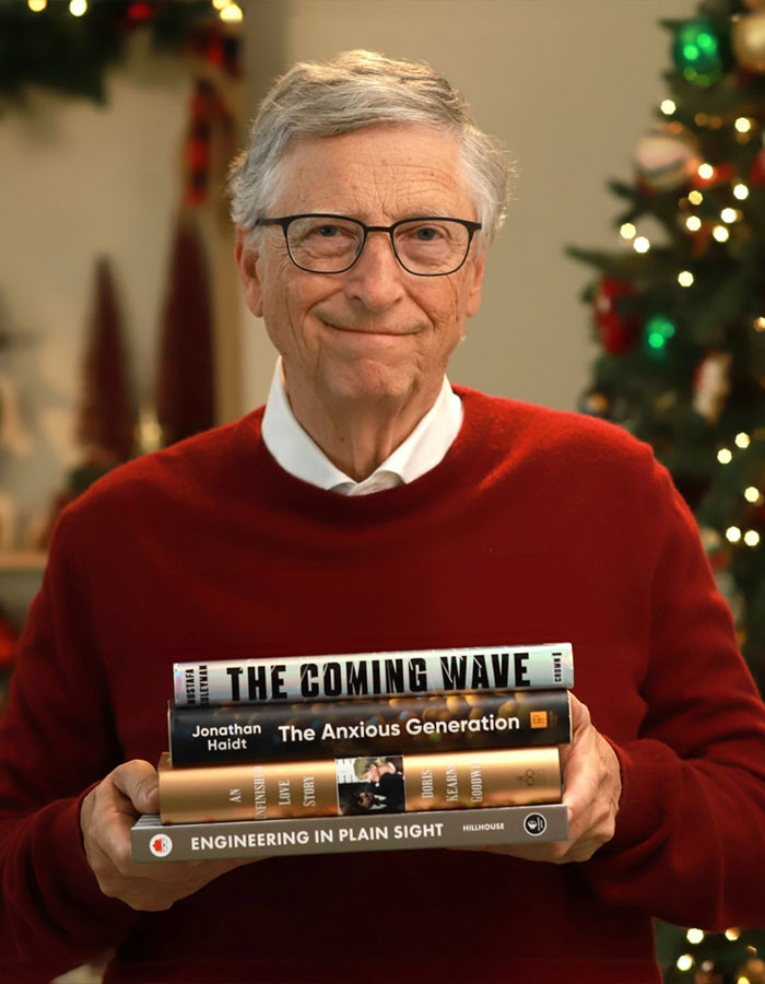 Man in glasses and red sweater holding a stack of books, with decorative lights in the background; relates to Epstein Island topic.