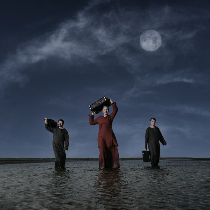 Three people in dark attire walk through water at night under a full moon, holding briefcases. Budapest International Foto Awards.