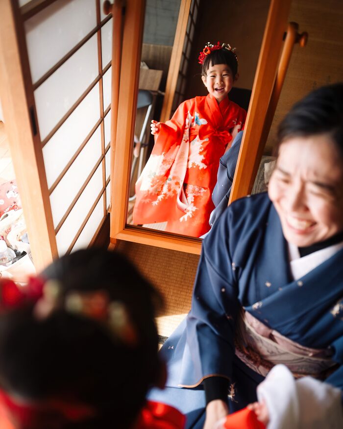 Child in a red kimono smiles in a mirror reflection, celebrating Budapest International Foto Awards 2024.