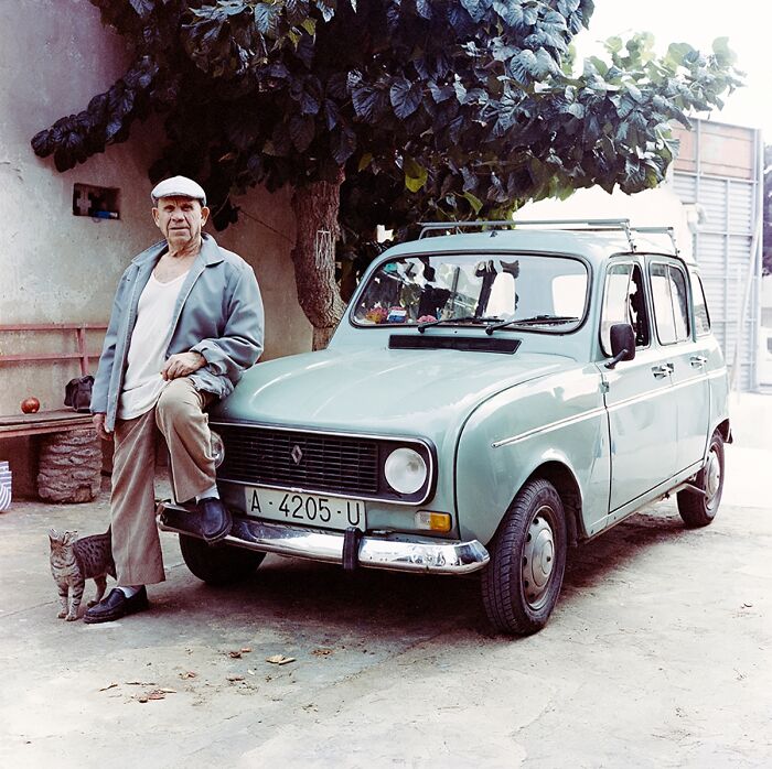 Man in a cap leans on a vintage car with a cat nearby, showcasing photography from Budapest International Foto Awards.