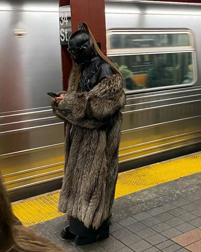 A funny subway passenger in a full costume and fur coat using a phone on the platform.