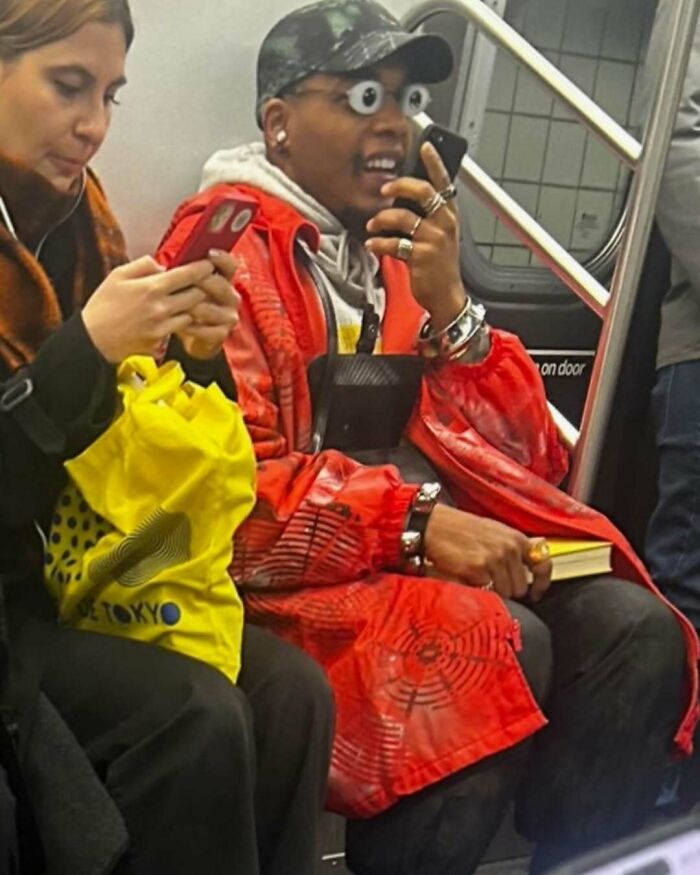 A NYC local in a bold red jacket and googly-eye glasses rides the subway, showcasing experimental fashion.