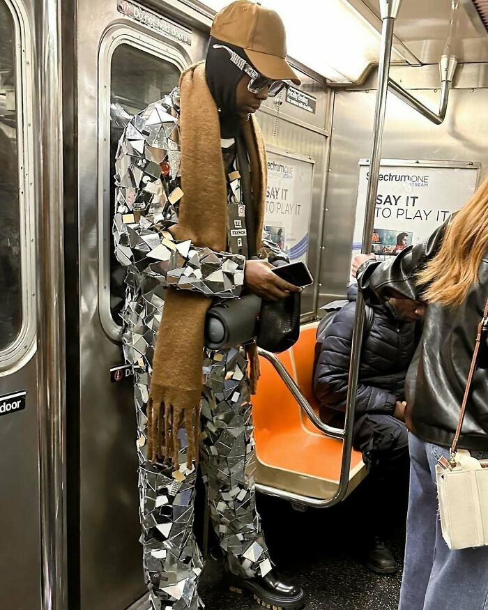 NYC local in a mirrored suit and cap, holding a phone on a subway, showcasing bold fashion experimentation.