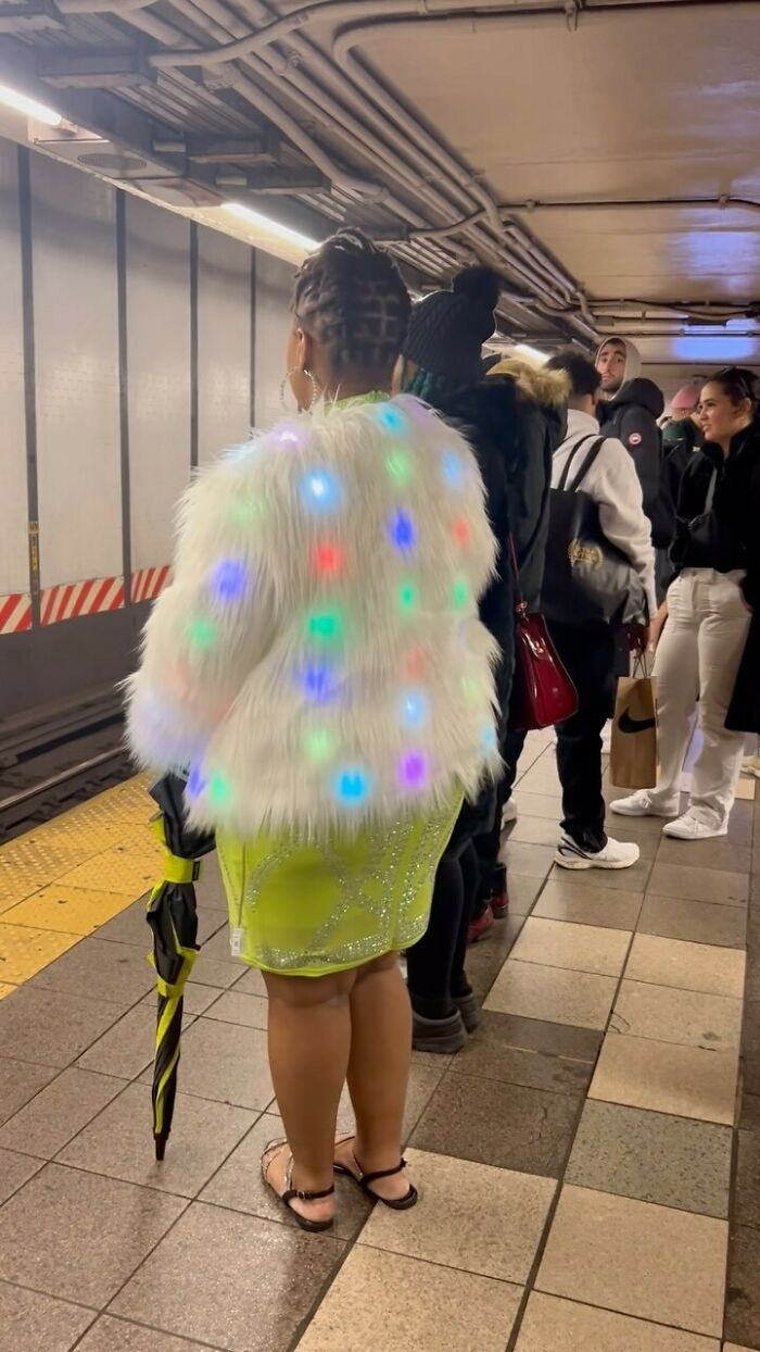 NYC local wearing a colorful, light-up fur jacket and neon dress standing on a subway platform, showcasing bold fashion.