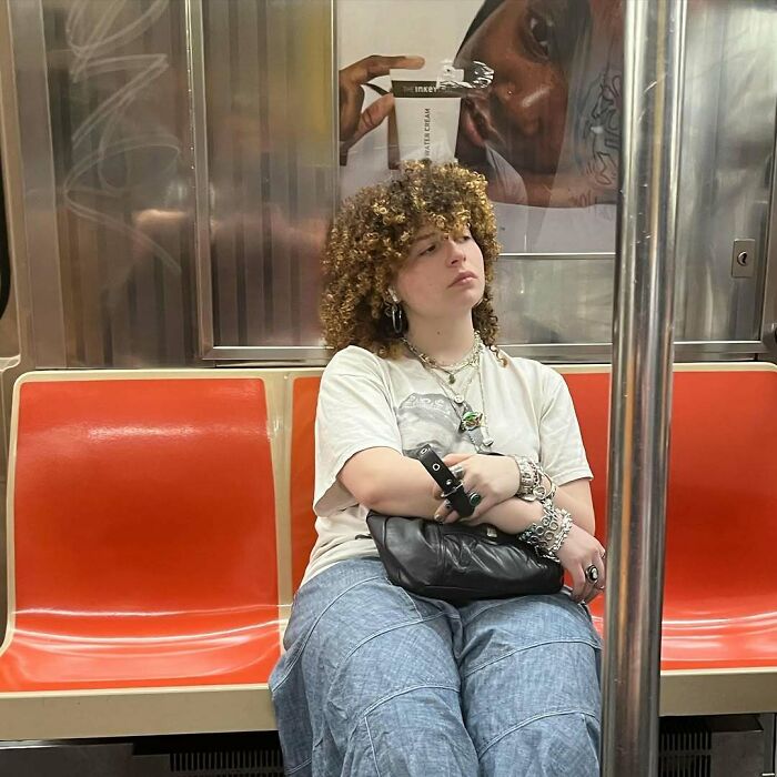 Curly-haired subway passenger with jewelry and a unique style sits on a train, emphasizing funny train experiences.