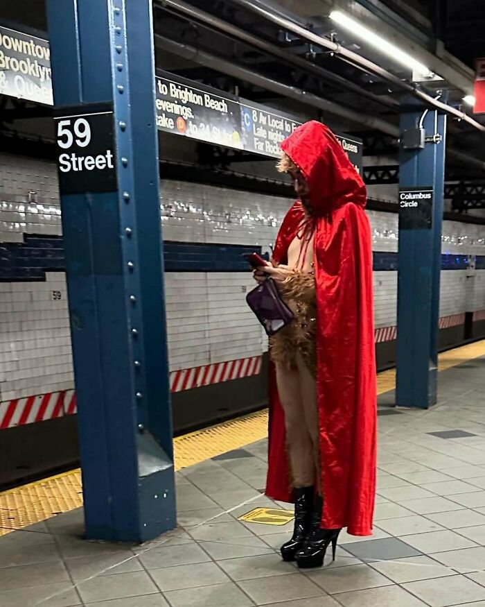 NYC local wearing a bold red hooded cape and furry attire at 59th Street subway station, experimenting with fashion.