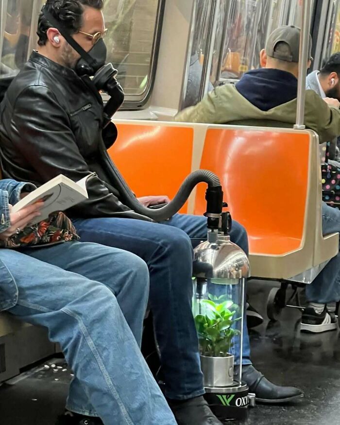 Man on subway with funny DIY air filter mask connected to a plant in a clear container on the floor.