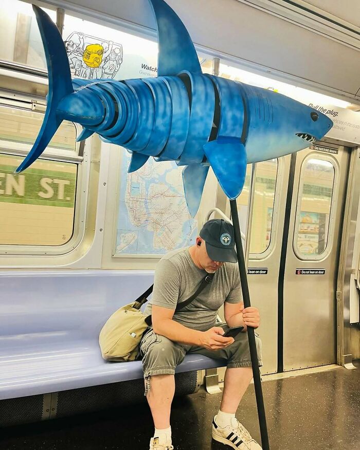 Man on subway wearing cap, holding a large blue shark sculpture, sitting on a bench, using phone. Funny train subway scene.