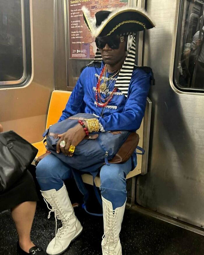 NYC local in a bold blue outfit with pirate hat, sunglasses, and boots, showcasing experimental fashion on the subway.