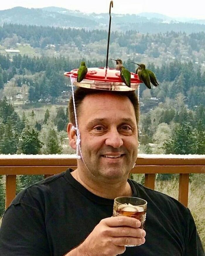 Man with a bizarre hummingbird feeder hat holding a drink, standing in front of a scenic landscape.