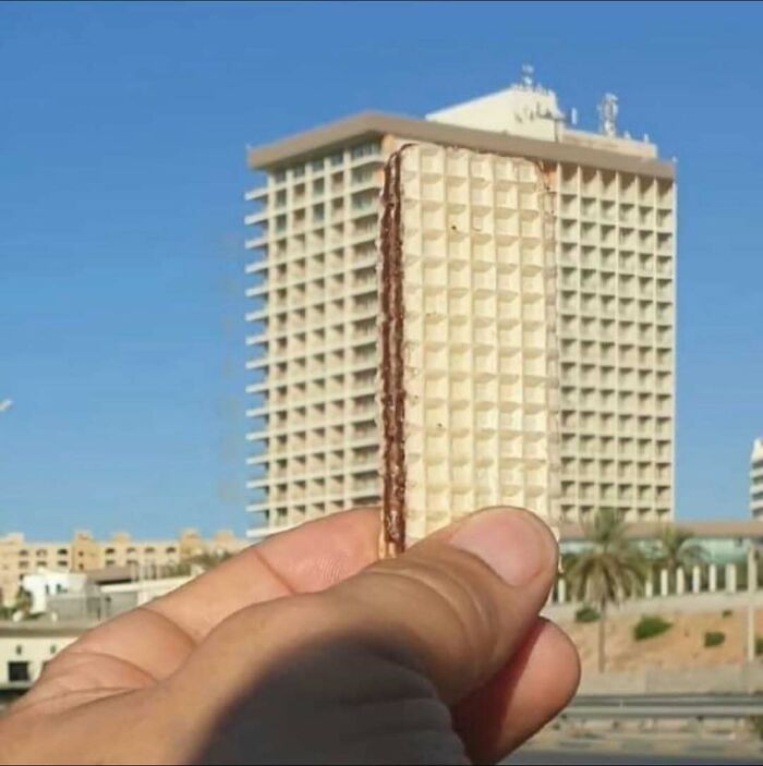 Hand holding a wafer in front of a building, creating a visual illusion on a bizarre Instagram page.