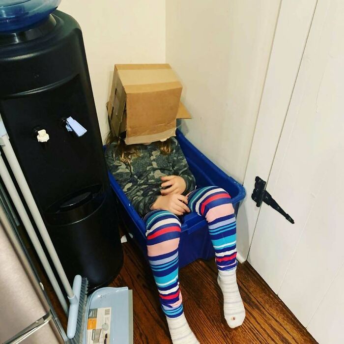 Child with a cardboard box on their head sits humorously in a blue bin, wearing colorful striped pants. Hilarious kids scene.