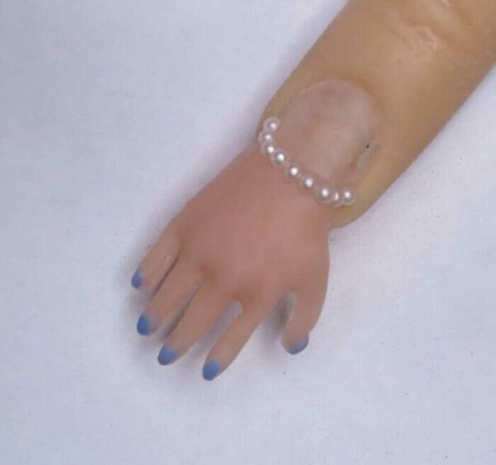 Bizarre photo of an unusual doll hand with blue nails and a pearl bracelet on a white background.